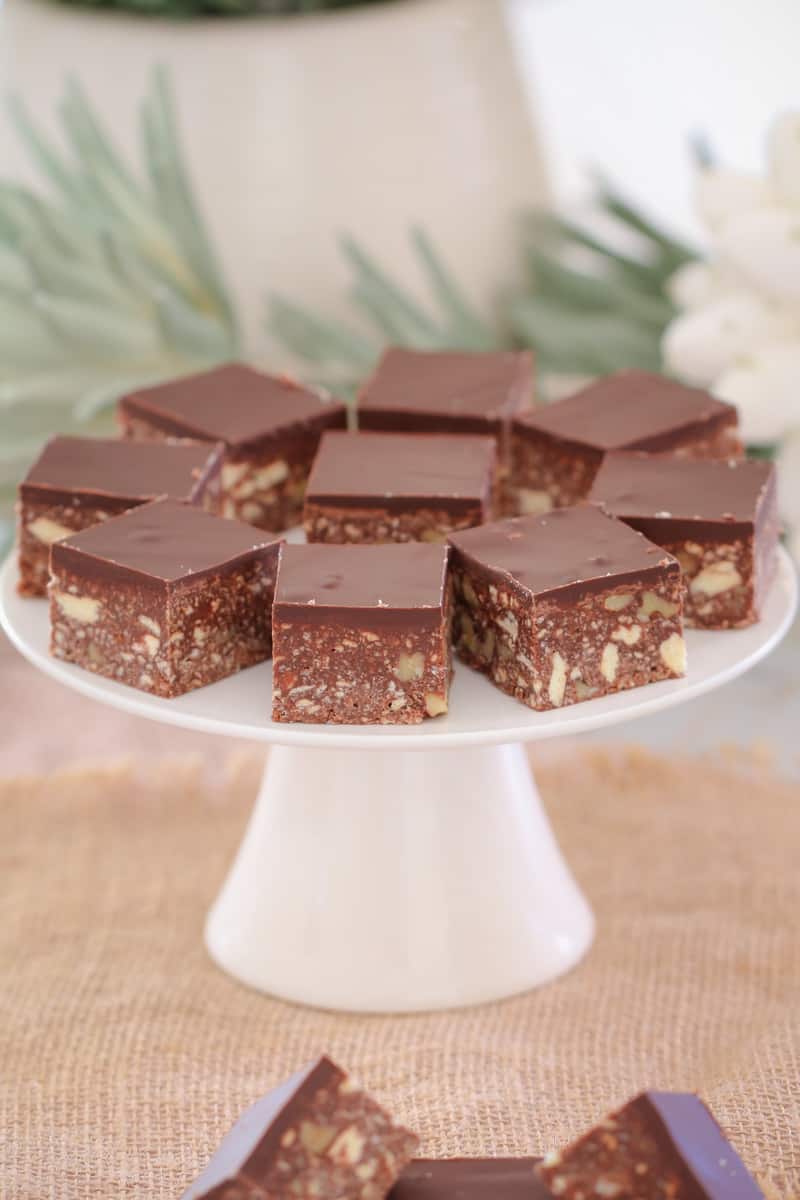 Squares of hedgehog slice with nuts and biscuits on a white cake stand.