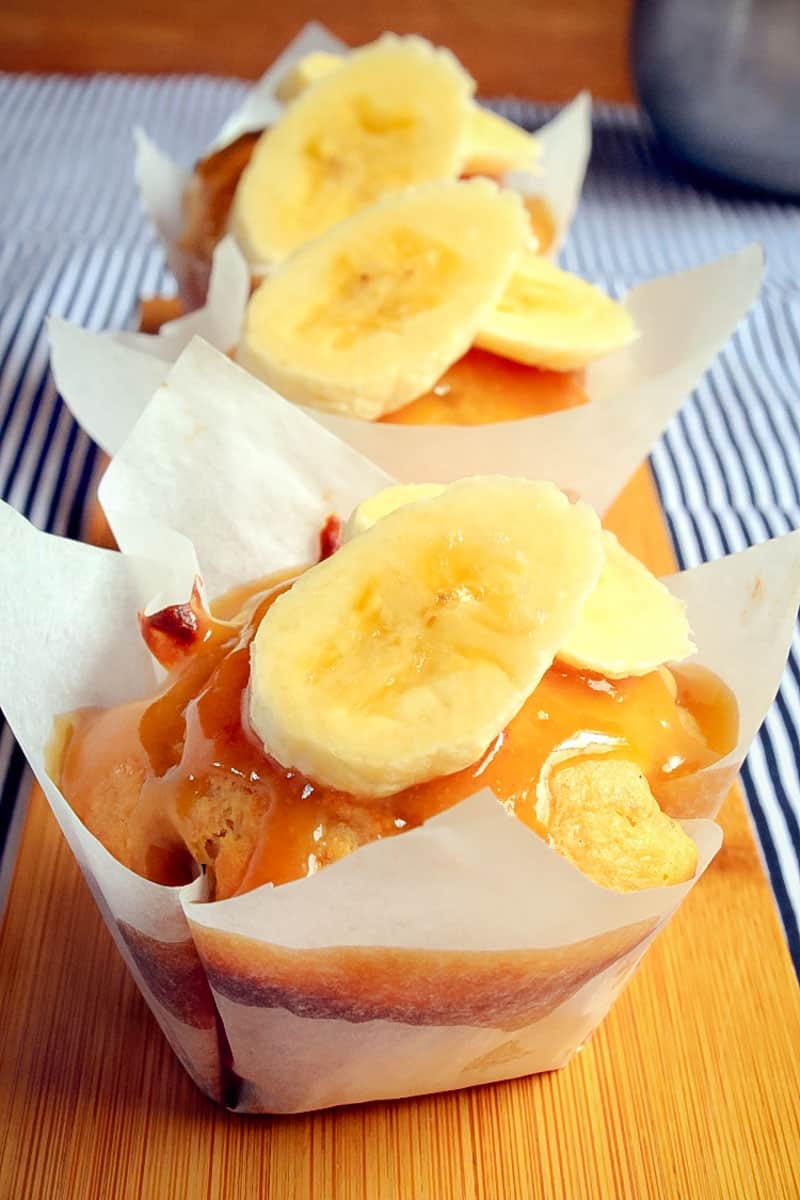 Two muffins on a board, each wrapped in baking paper, drizzled with a caramel sauce and topped with banana slices