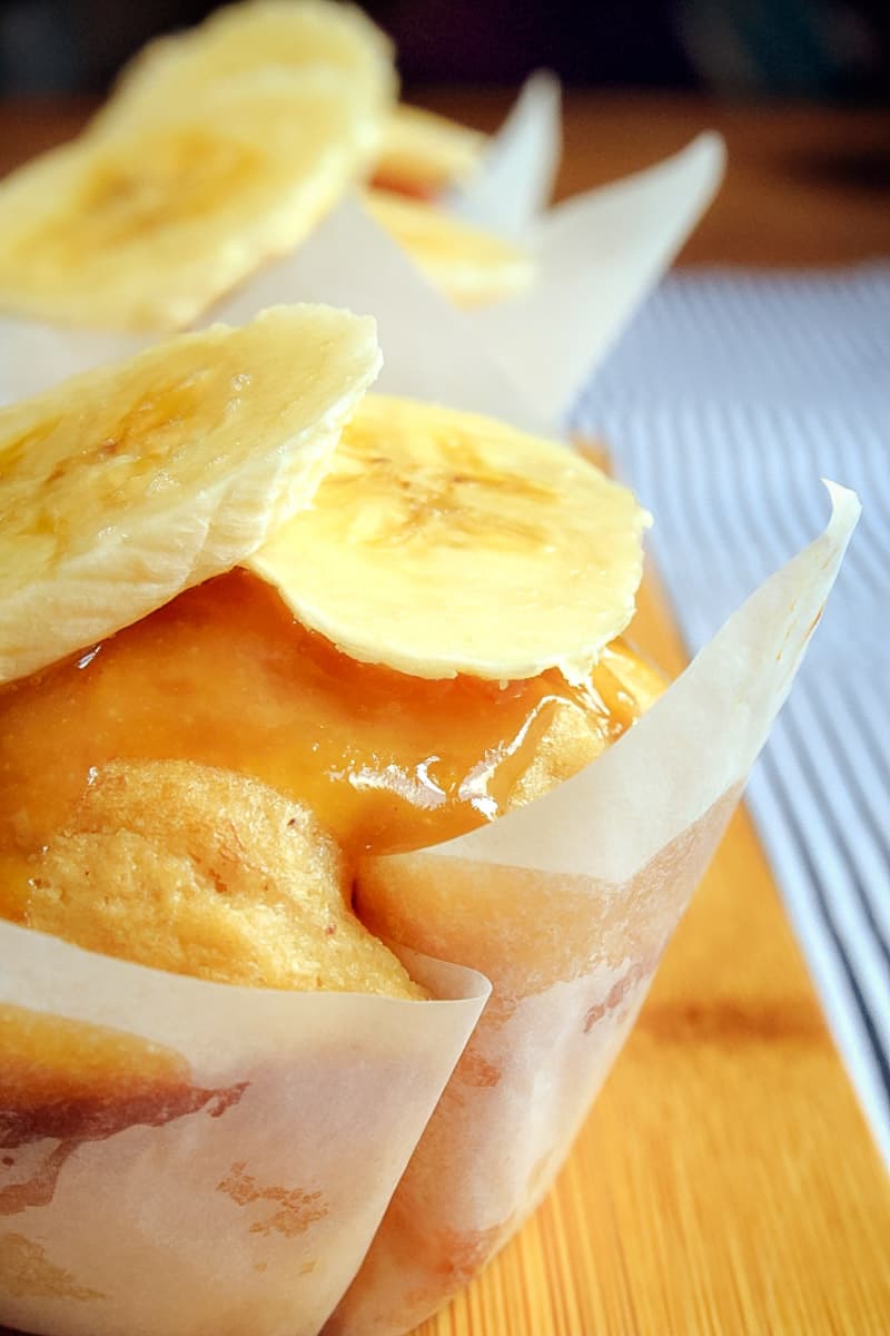 A close up a muffin wrapped in baking paper, drizzled with caramel sauce and topped with banana slices