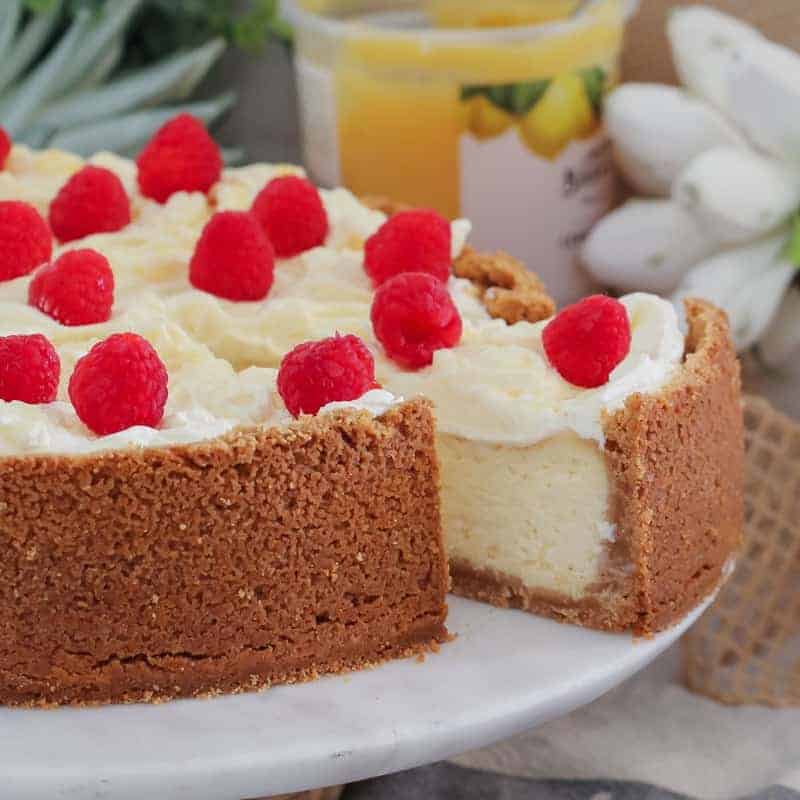 The ultimate classic Baked Lemon Cheesecake... a delicious biscuit base filled with creamy, tangy lemon cheesecake and topped with whipped cream, swirls of lemon curd and fresh raspberries. 