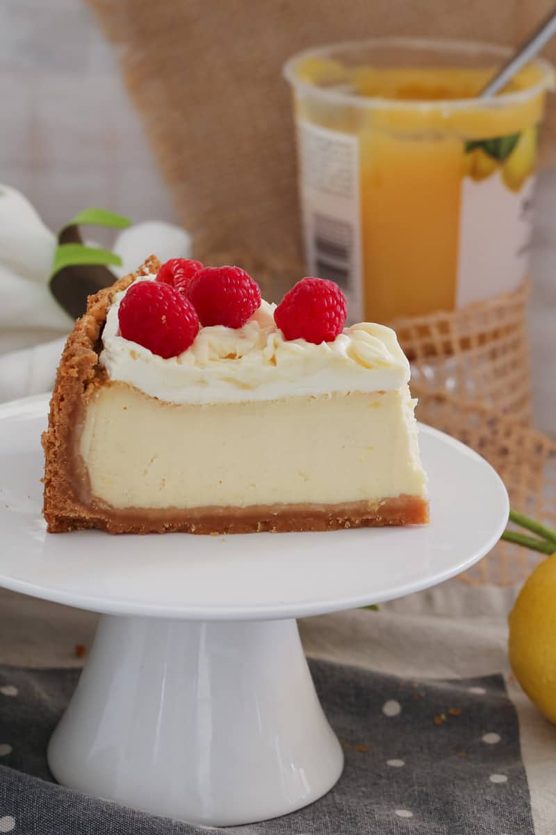 A side view of a cut slice of cheesecake topped with whipped cream and fresh raspberries on a white cake stand