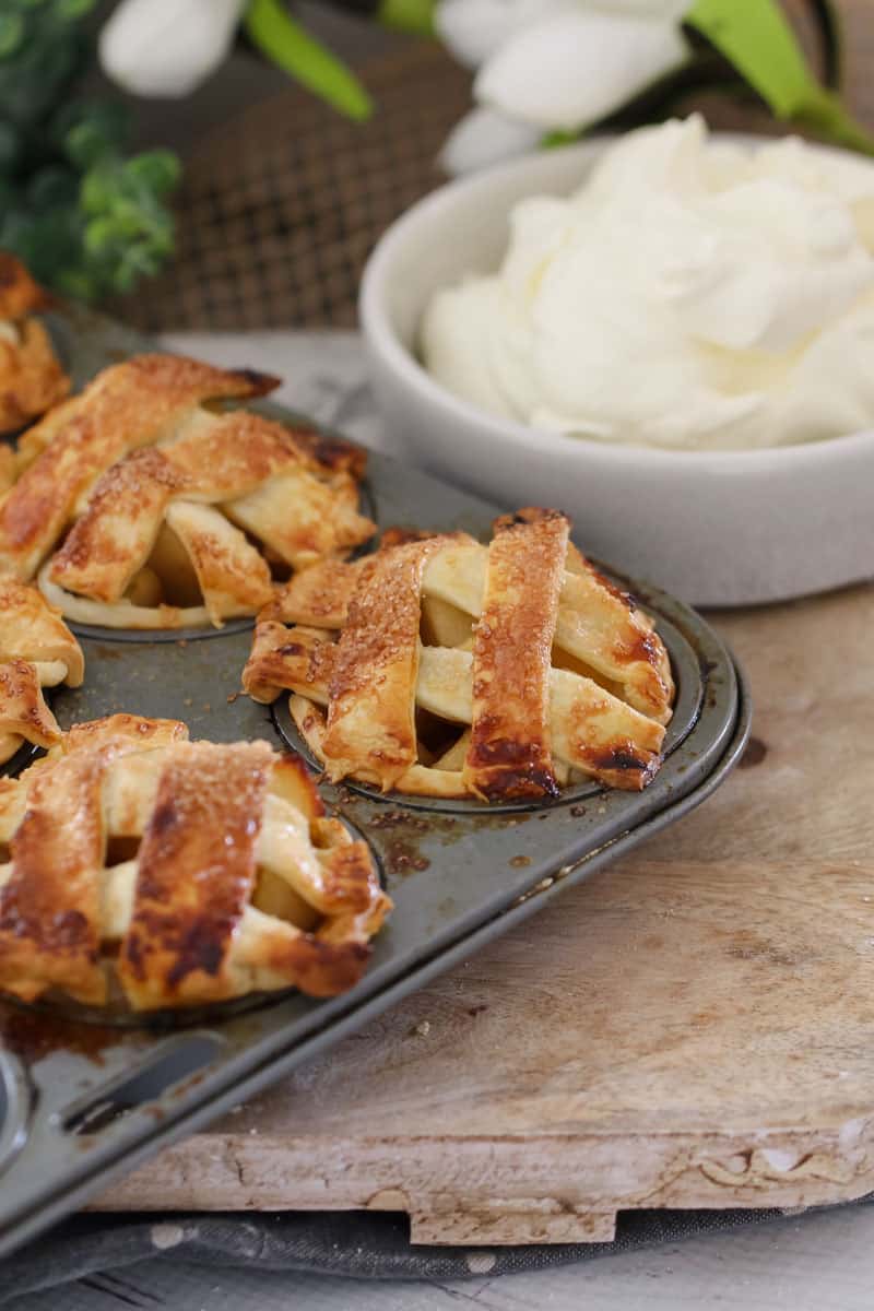 A muffin tray filled with mini apple pies topped with strips of pastry and a bowl of whipped cream