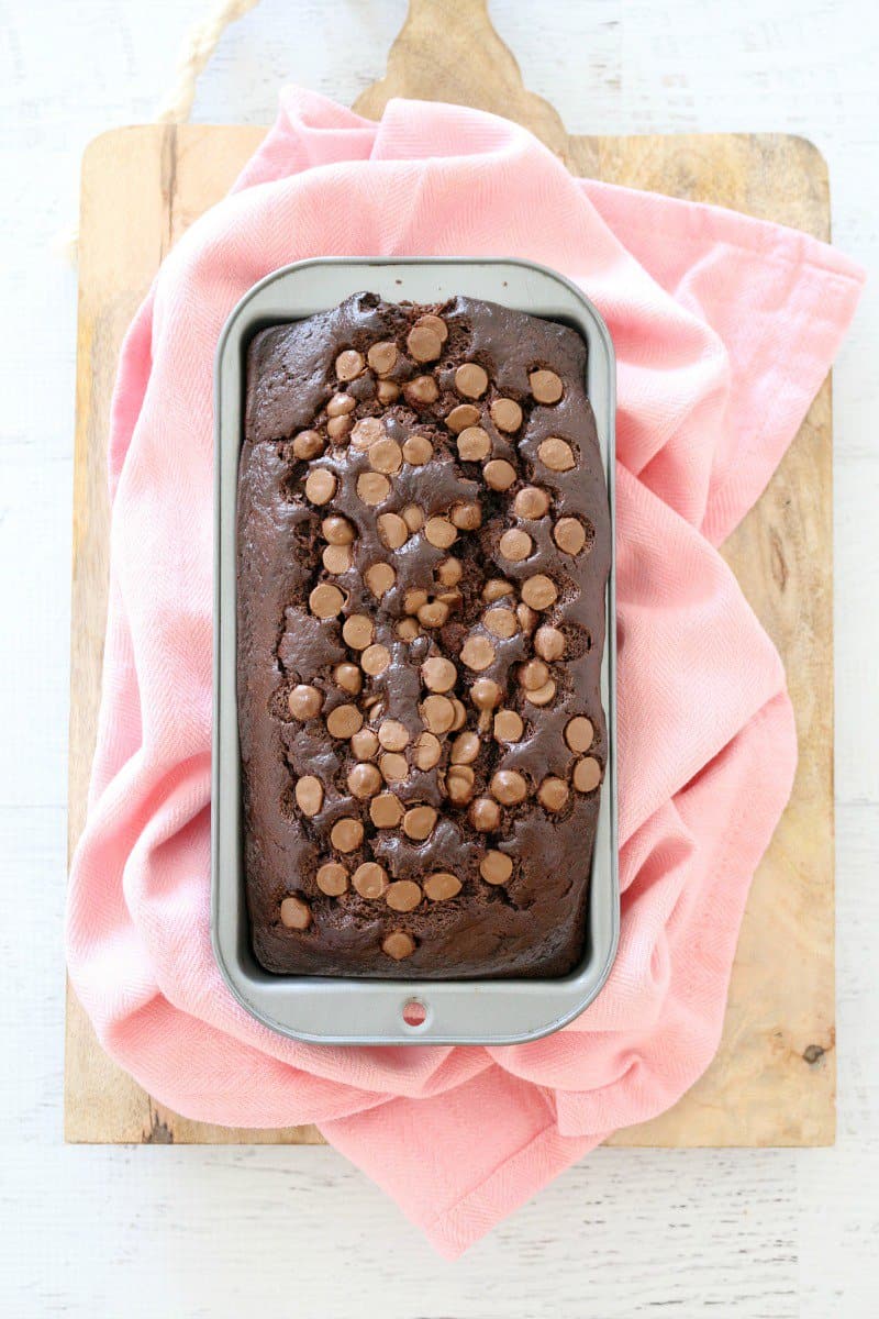 A baked banana and chocolate loaf covered in chocolate chips, in a loaf tin resting on a pink tea towel.