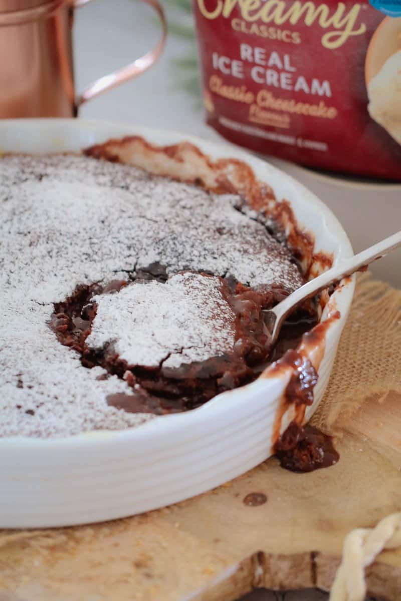 A white baking dish of chocolate pudding dusted with icing sugar with a spoonful showing chocolate sauce inside
