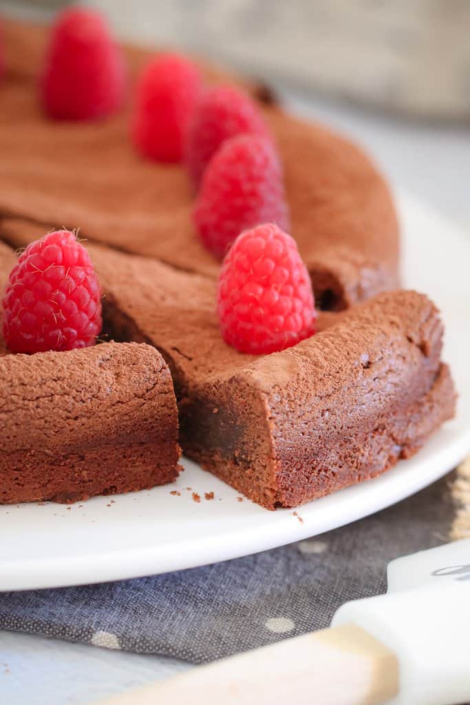 A chocolate torte decorated with fresh raspberries on a plate with one serve cut and slightly removed