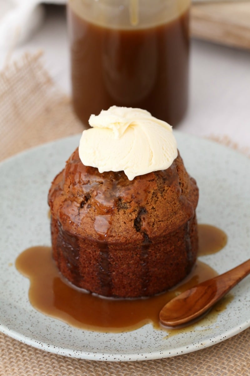 A mini sticky date pudding drizzled with caramel sauce and topped with whipped cream on a white plate