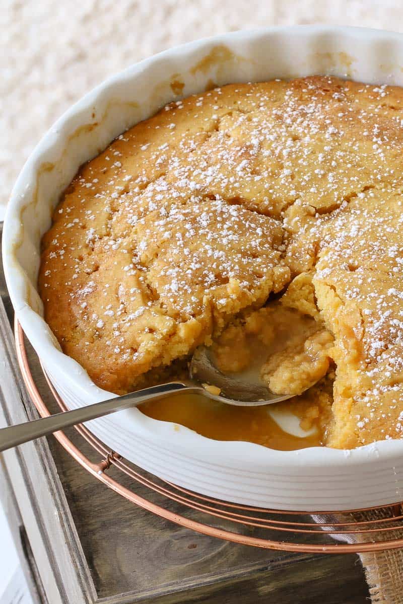 A round white baking dish of pudding dusted with icing sugar and a spoonful removed to show the sauce underneath