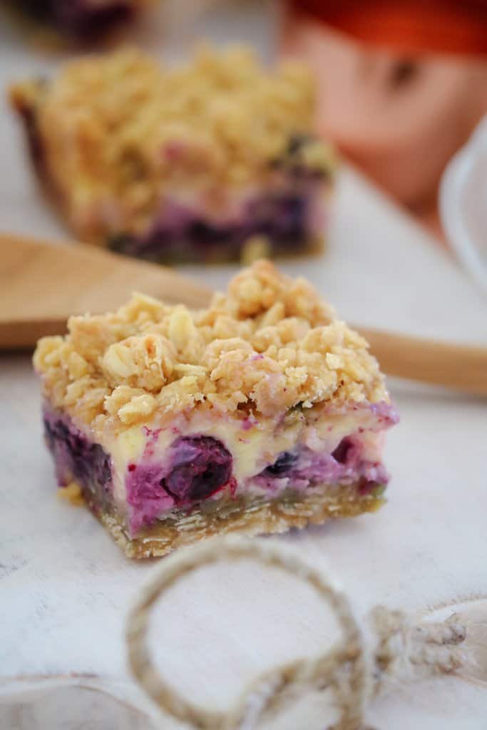 A square of slice with a crumble topping and filled with blueberries in front of a wooden spoon