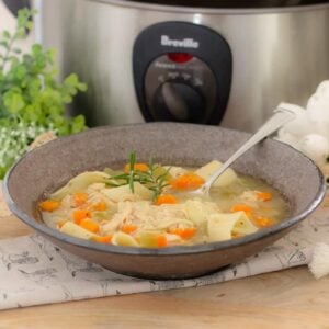 A bowl of chicken noodle soup in front of a slow cooker and herbs.