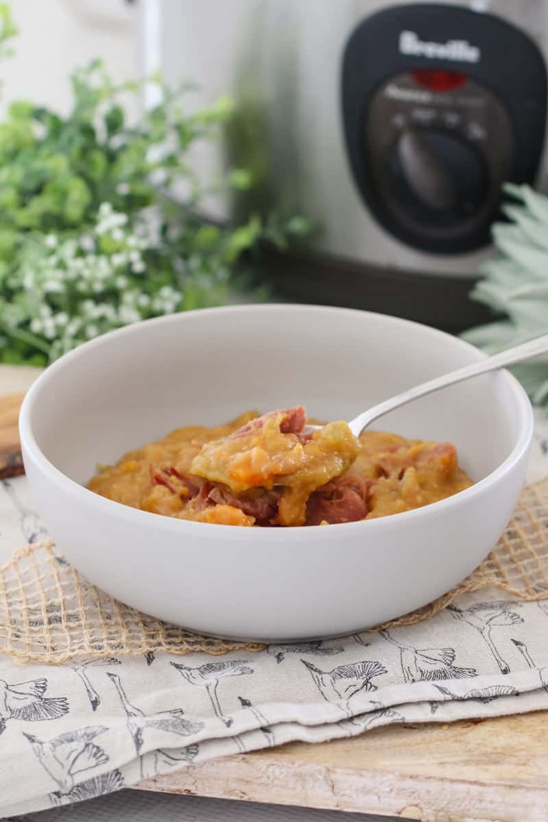 A spoon in a white bowl of yellow split pea soup with ham, in front of a slow cooker