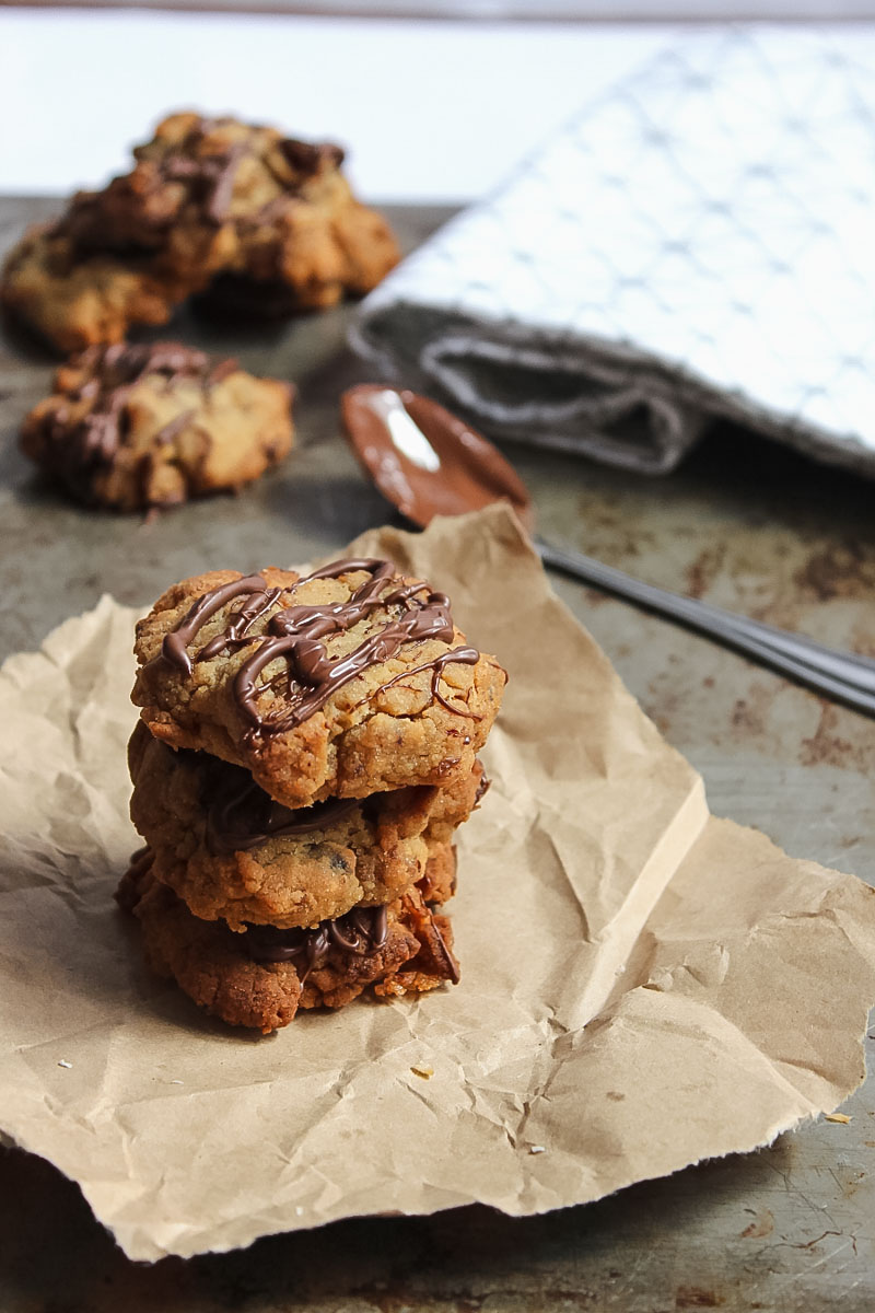 A stack of cookies filled with dates and drizzled with chocolate on a piece of paper beside a chocolate coated spoon
