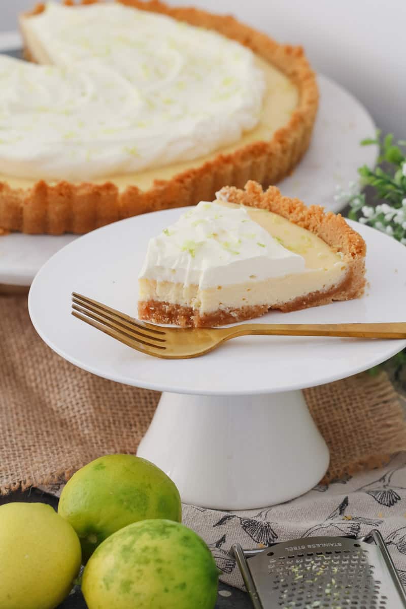 A piece of half eaten key lime pie on a plate next to a white cake stand with the rest of the pie.