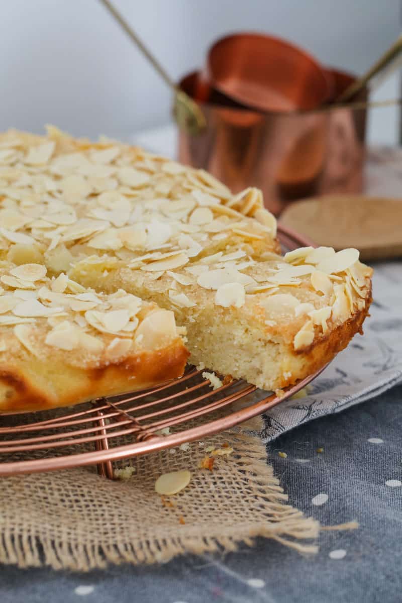 A lemon and ricotta cake topped with flaked almonds, on a copper wire tray, in front of copper measuring cups