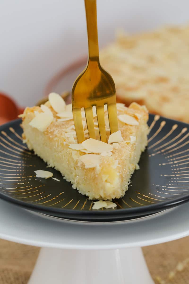 A piece of lemon cake topped with flaked almonds on a black plate with a gold fork standing up in it