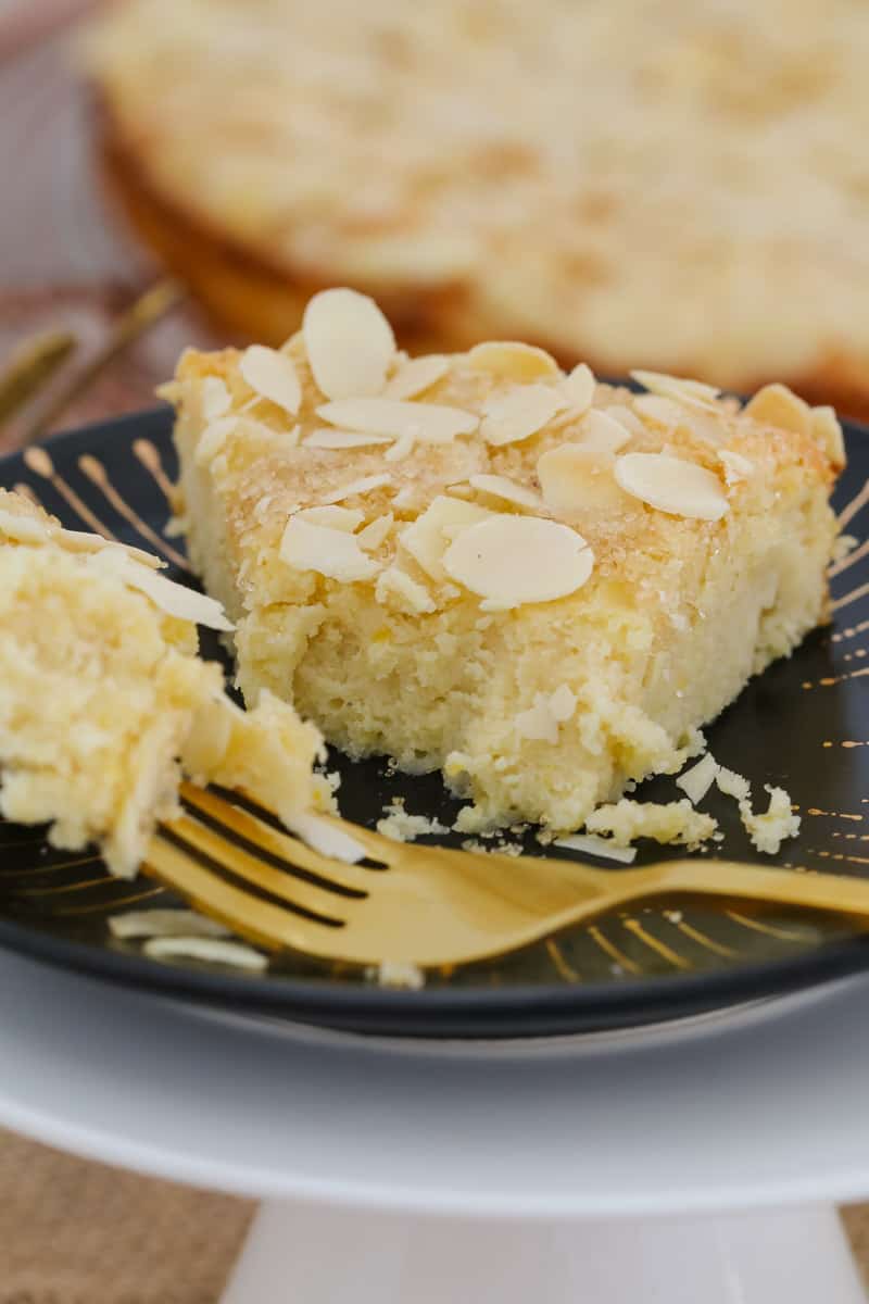 A close up of a slice of moist lemon and ricotta cake served on a black plate with a gold fork