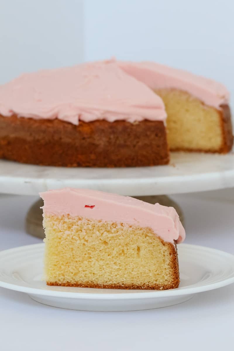 A piece of white cake covered in pale pink buttercream on a white plate.