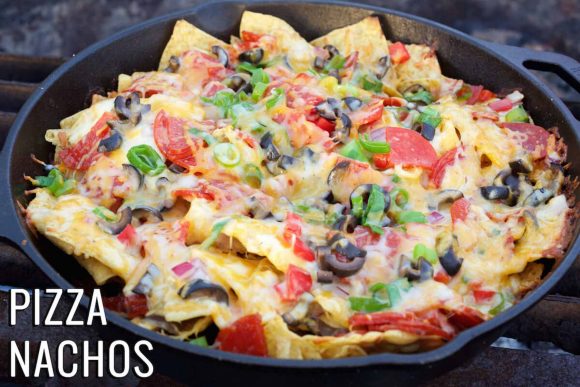 Nachos being cooked on a campfire with corn chips, chillies, olives, tomato and cheese. 