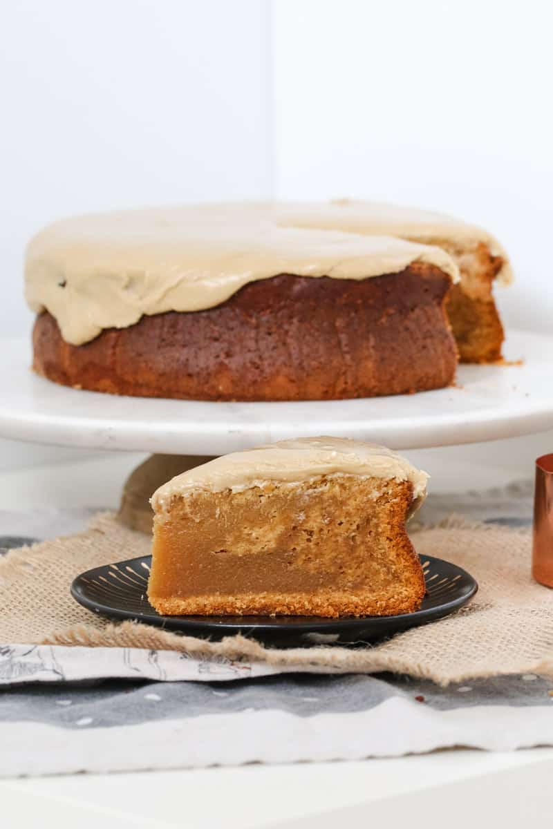 A serve of caramel mud cake on a black plate, in front of the remaining cake on a cake stand