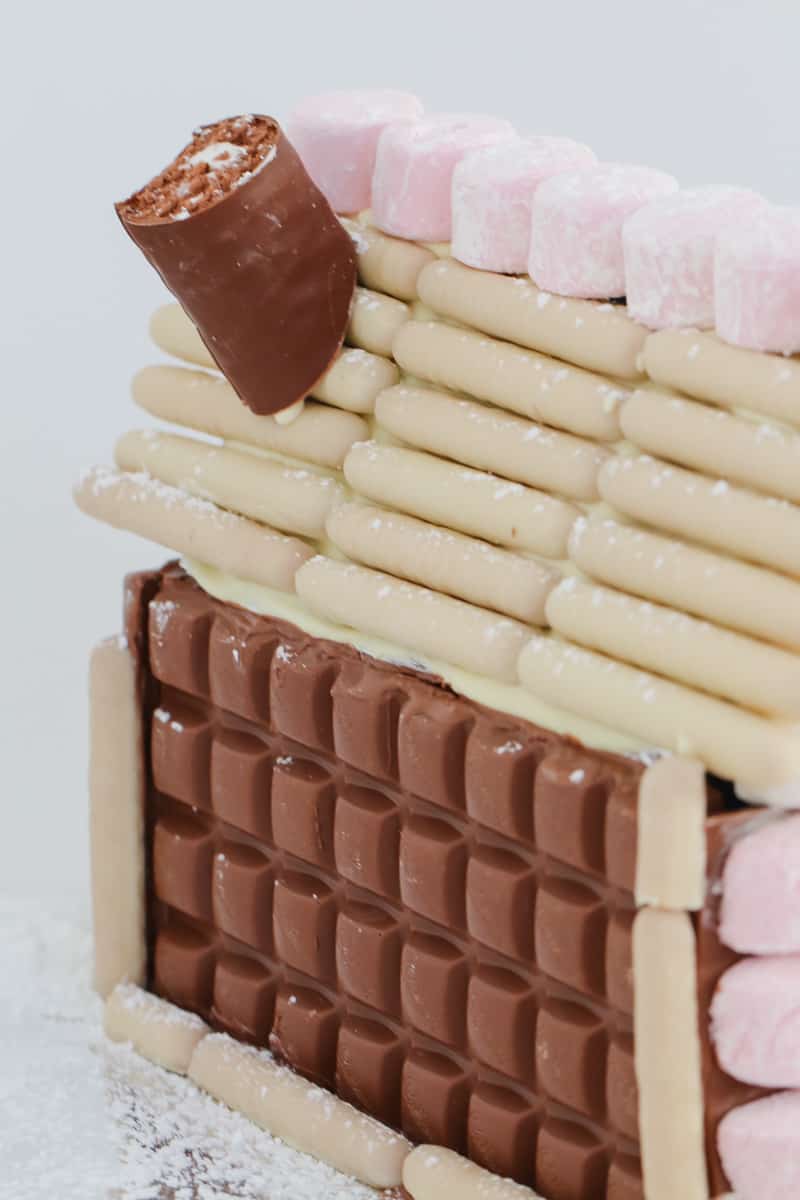 A close up of the side of a gingerbread house, decorated with chocolate bars, chocolate logs and marshmallows.