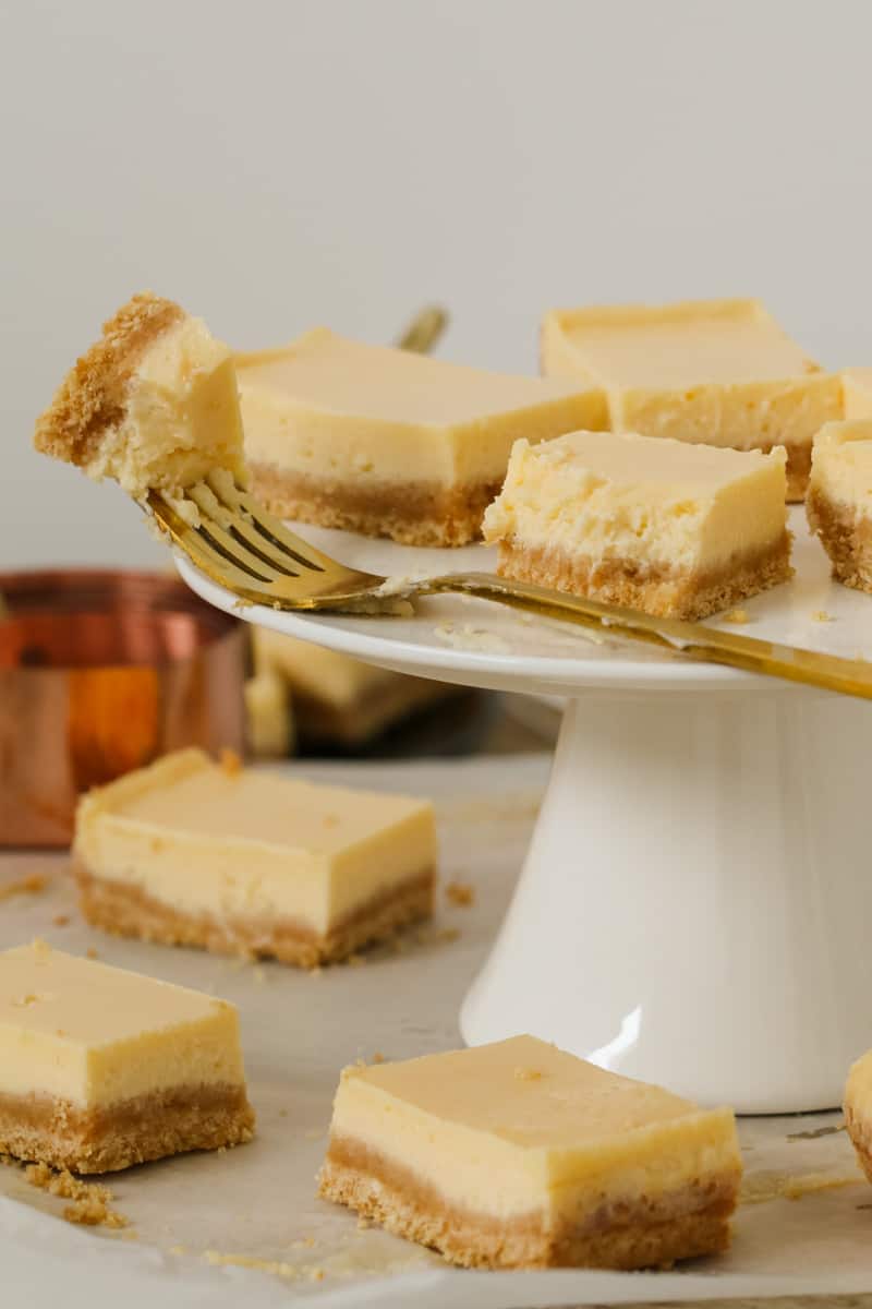 A gold fork holding a piece of creamy lemon slice and resting on a white cake stand with more squares of slice