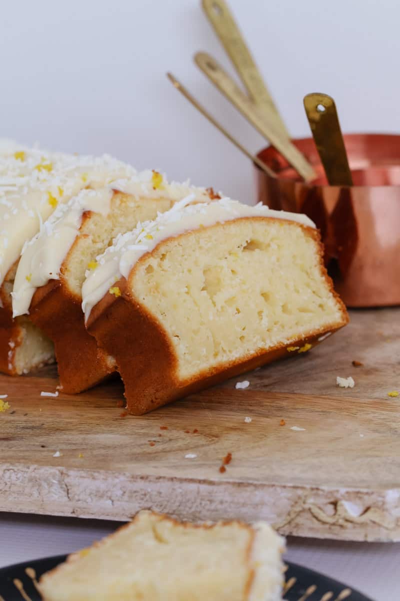 Slices of iced lemon coconut loaf on a wooden board in front of a copper measuring cup