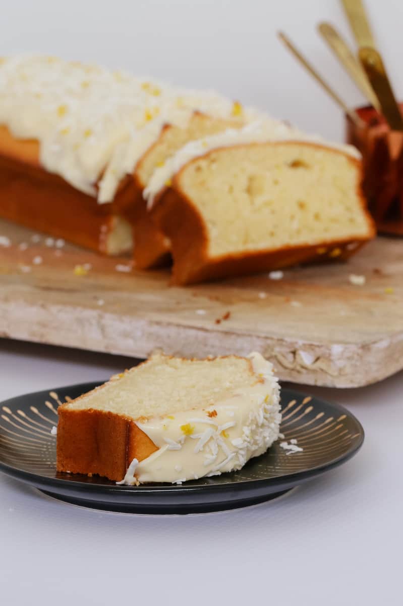 A slice of iced lemon coconut loaf on a black plate, in front of a wooden board with more slices.
