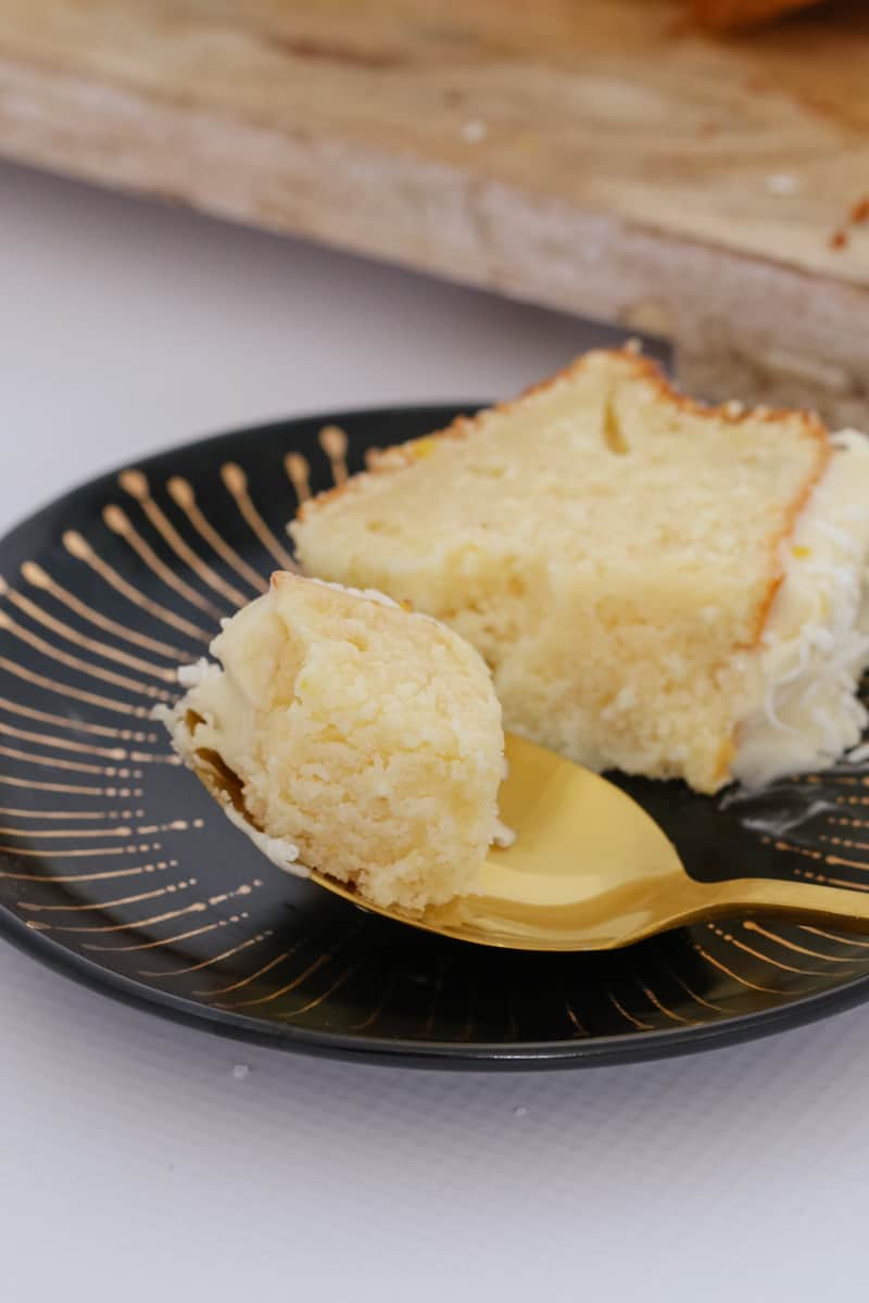 A slice of iced lemon coconut loaf on a black plate with a spoonful on a gold spoon