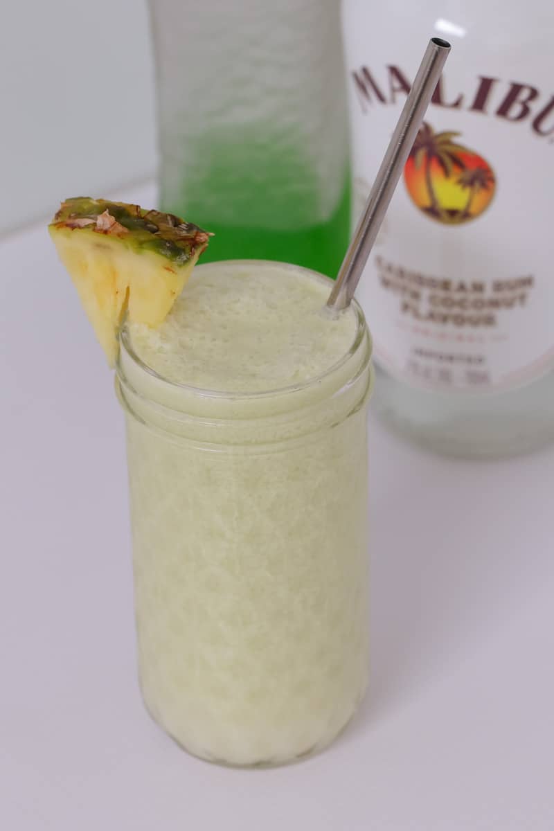A glass jar filled with a creamy cocktail and pineapple wedge in front of bottles of Midori and Malibu