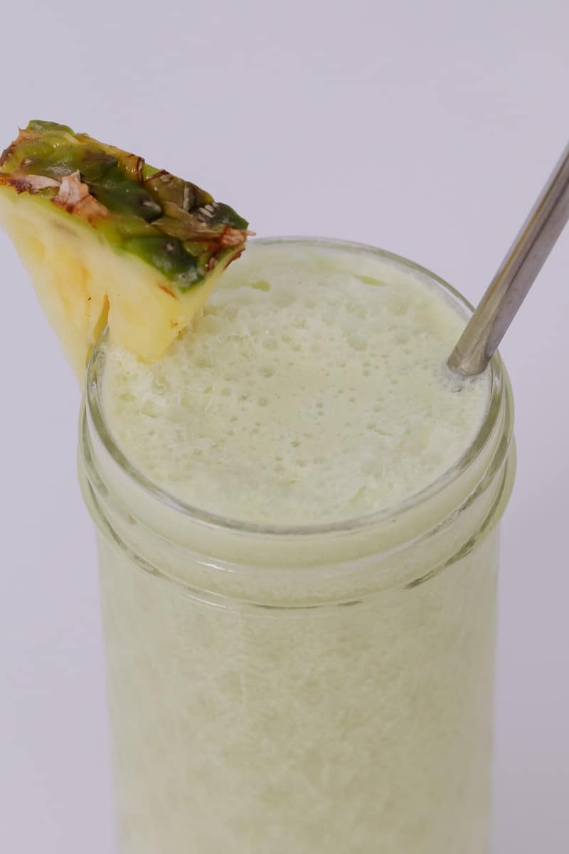 Looking down at a textured glass jar filled with a cream coloured cocktail, a wedge of pineapple and a stainless straw