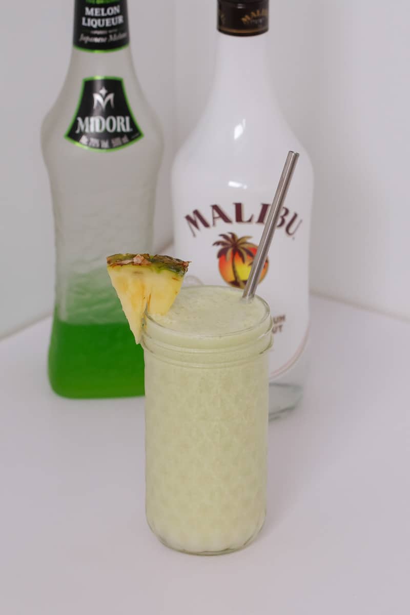 A textured glass jar filled with a creamy cocktail in front of bottles of Midori and Malibu