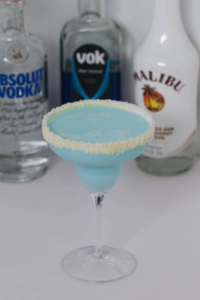 A blue cocktail in a sugar rimmed cocktail glass in front of bottles of vodka and Malibu