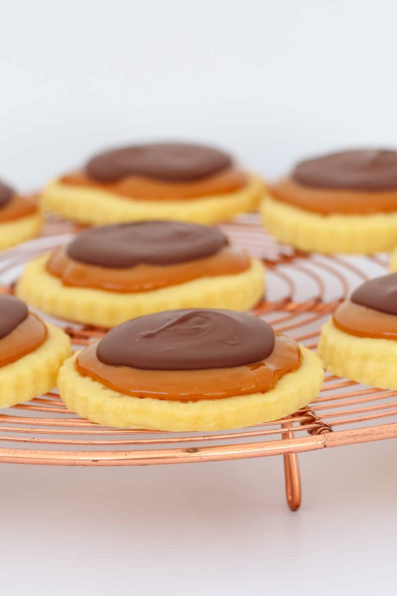 A copper wire rack topped with round shortbread cookies, thick caramel and melted chocolate. 