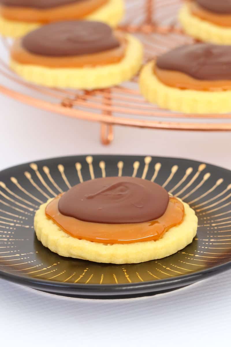 A cookie topped with caramel and chocolate on a black plate, in front of a copper wire stand with more cookies