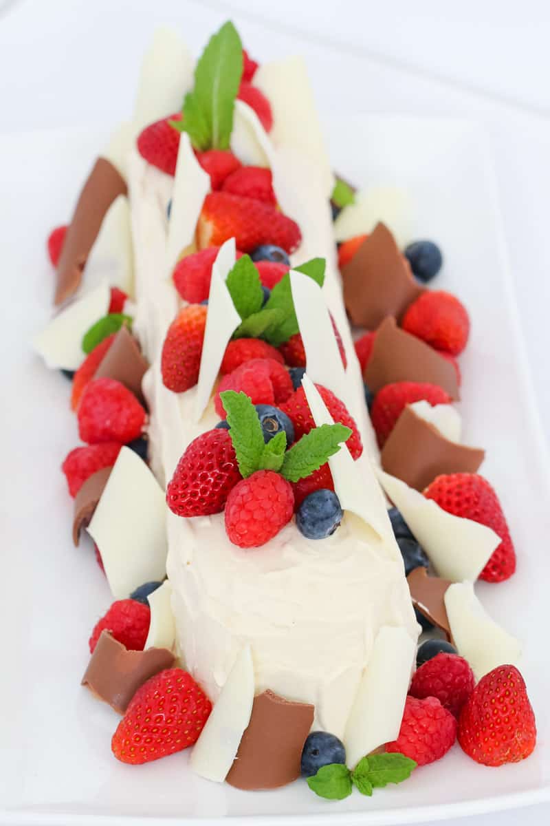 A close up of a cream covered log, decorated with fresh berries and white and milk chocolate shards