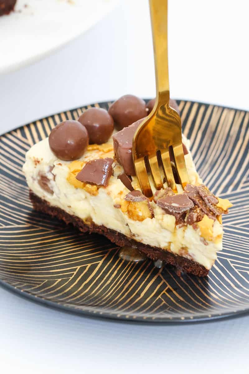 A gold fork being pressed into a slice of cheesecake topped with honeycomb and chocolate malted milk balls