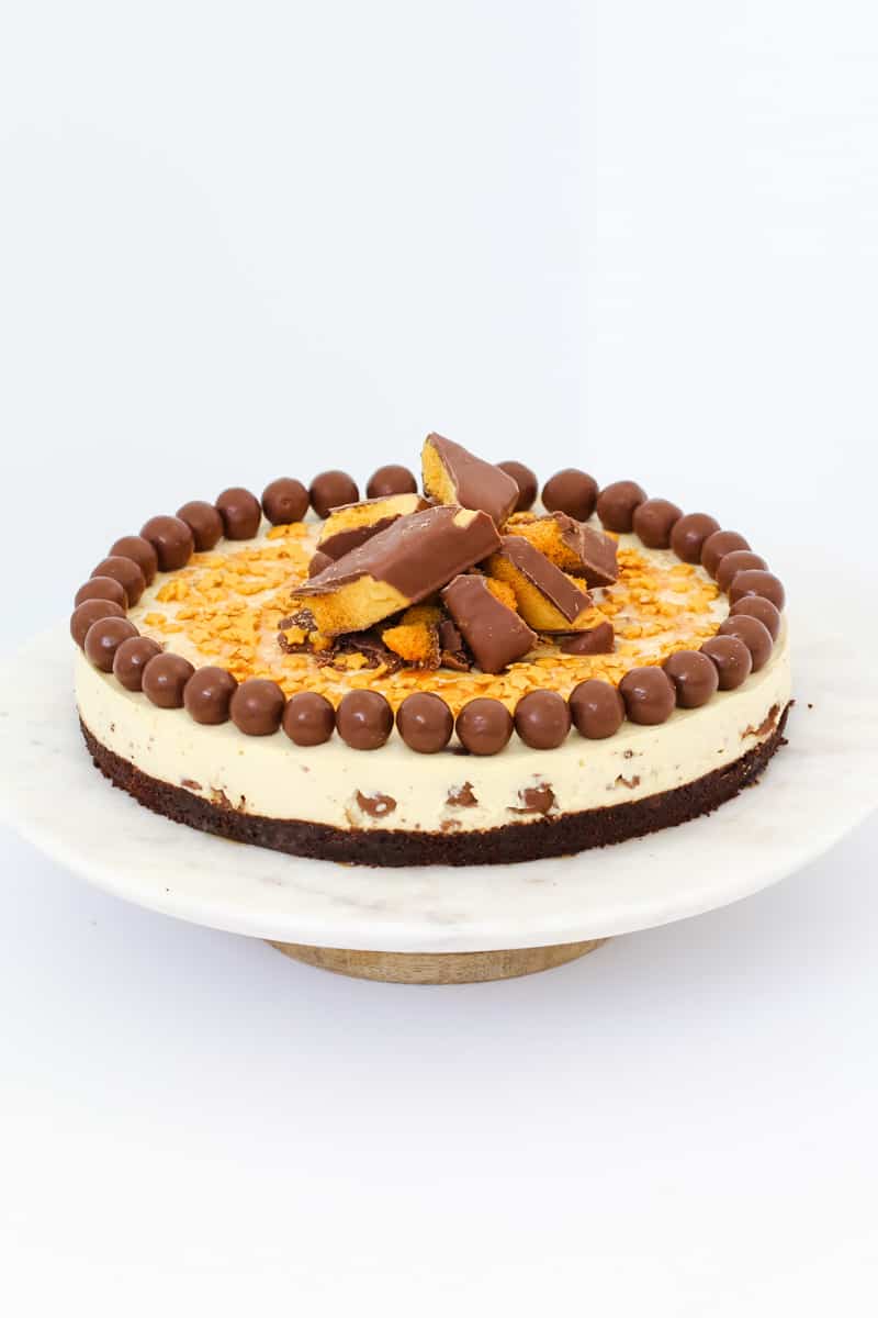 A round cheesecake with a chocolate base  and creamy filling topped with chopped honeycomb and Malteser malted milk balls