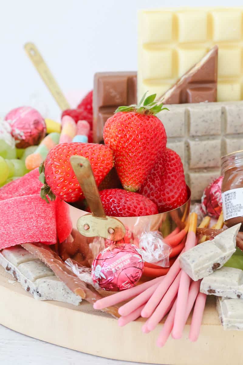 A close up of chocolate pieces, assorted lollies, and a copper measuring cup filled with strawberries.