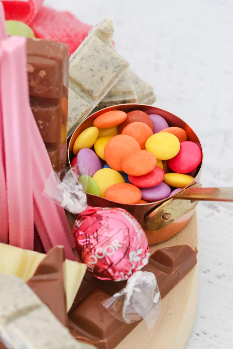 A close up of coloured Smarties in a copper measuring cup, surrounded by chocolate pieces and lollies
