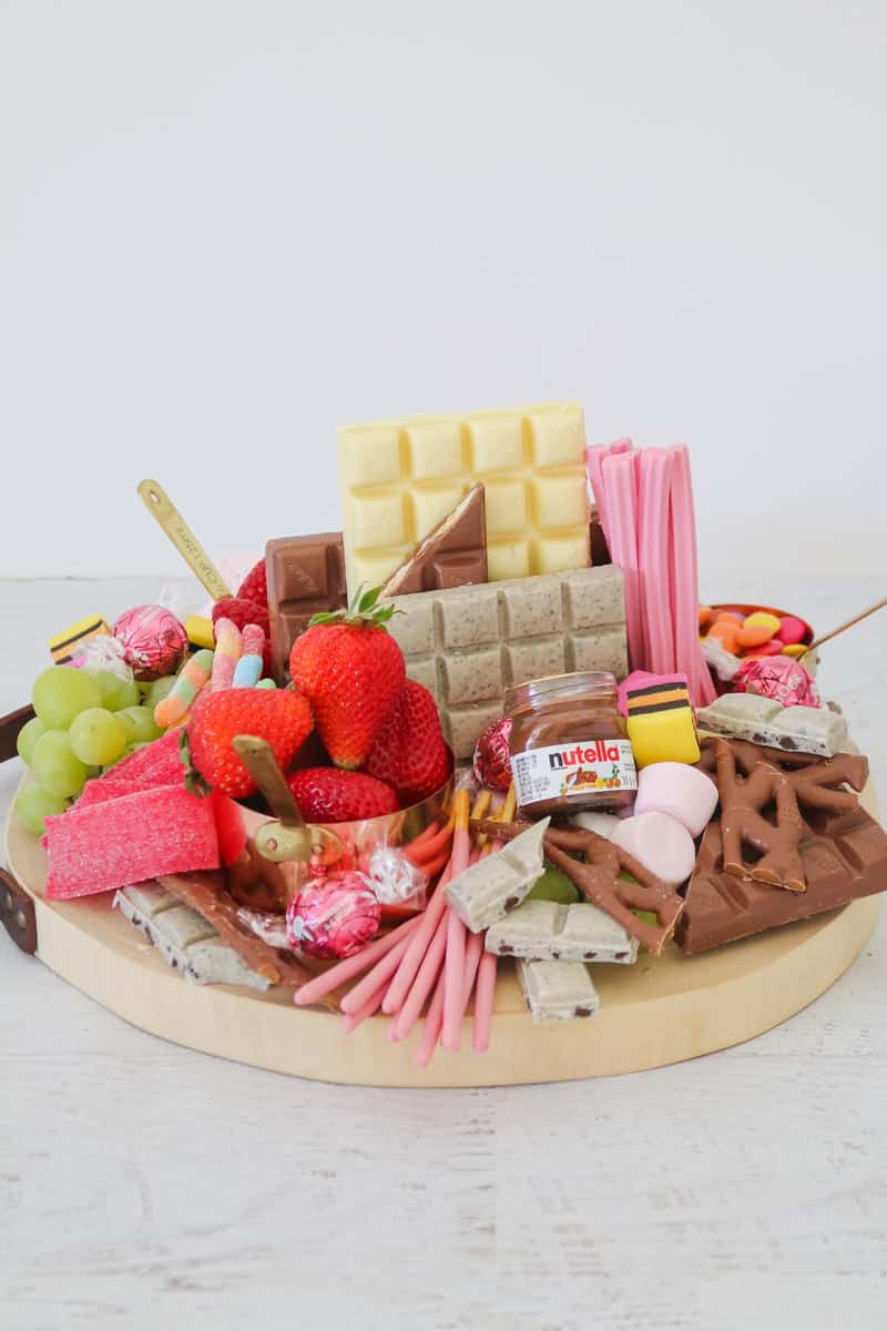 A round wooden board packed with chocolate bars, fruit, Nutella in a jar and assorted lollies.