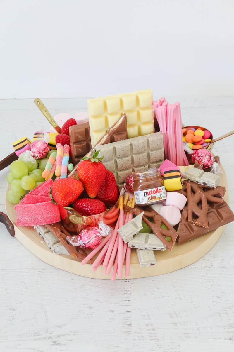 A round wooden board packed with chocolate bars, fruit and assorted lollies.