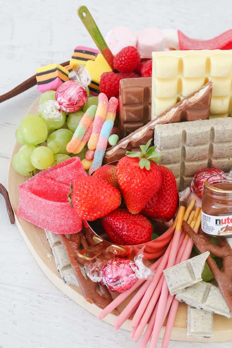 A close up of a wooden board packed with a selection of chocolate bars, grapes, strawberries, raspberries and assorted lollies.