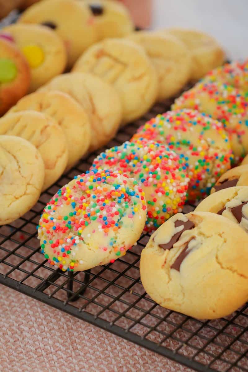 Three lines of cookies, one plain, one with coloured sprinkles, and one with chocolate chips on a wire tray