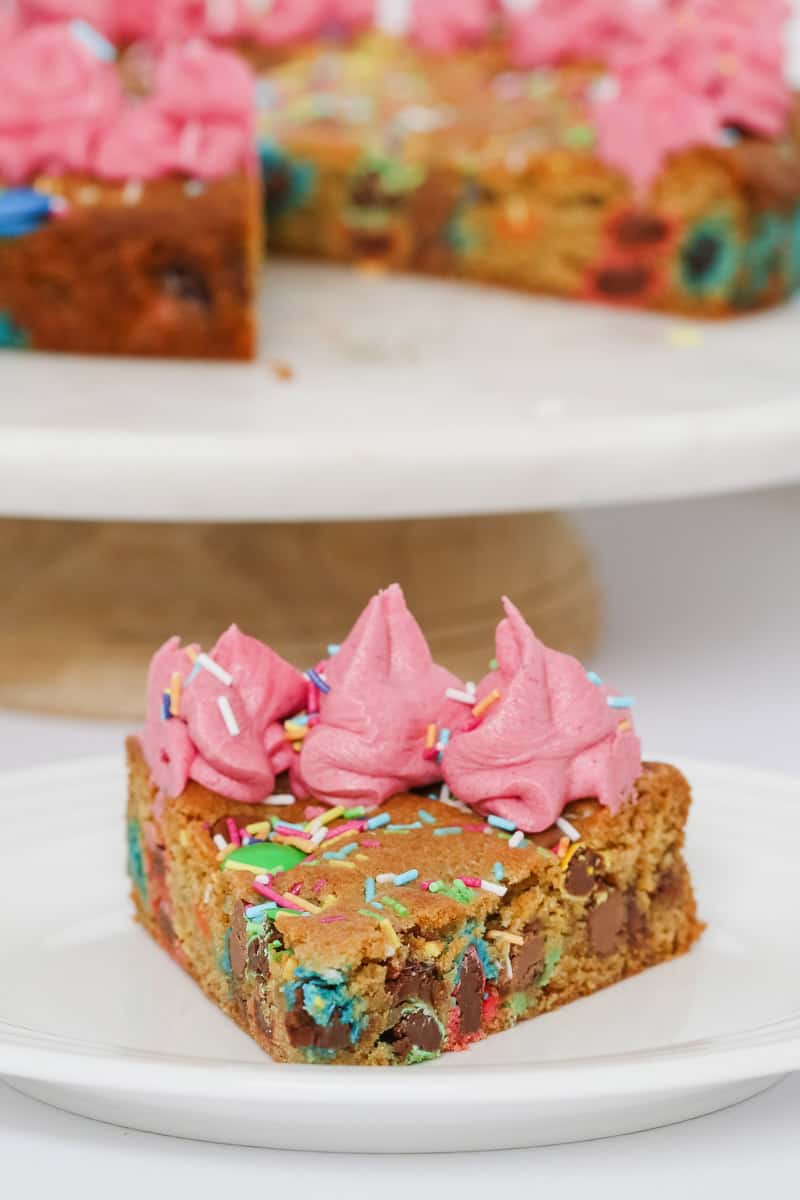 A slice of chocolate chip cookie cake with pink frosting and sprinkles on top, served on a white plate.