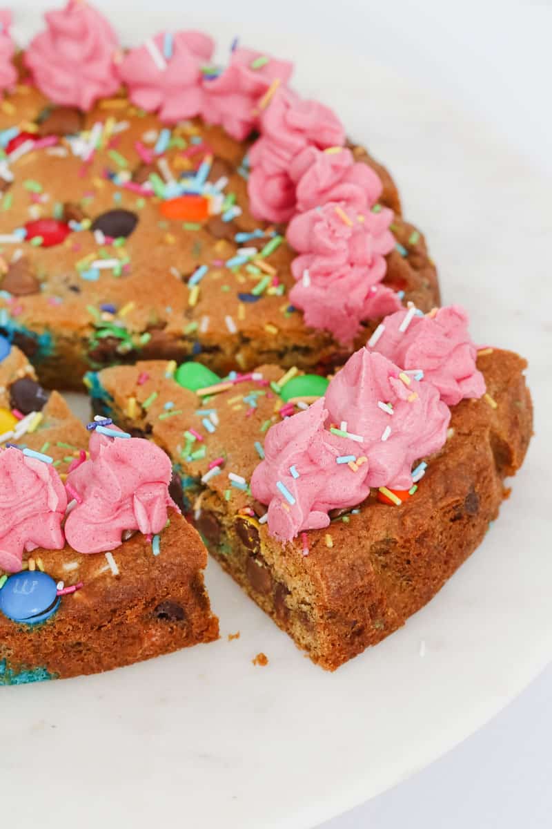 Slice of chocolate chip cookie cake with pink frosting and sprinkles on top, served on a white plate.