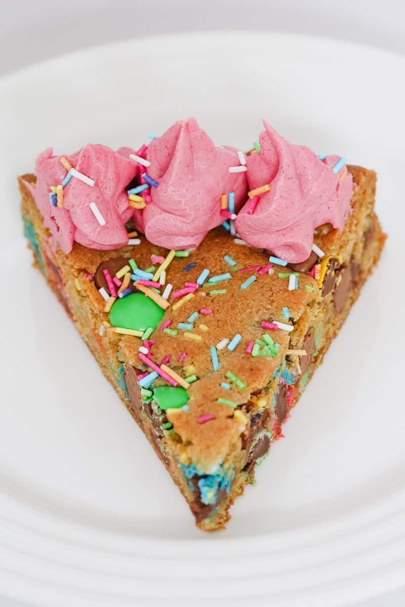 A slice of cake with M&Ms, chocolate chips, sprinkles, and pink buttercream frosting on a white plate.