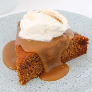 A slice of Sticky Date Pudding drizzled with caramel sauce and topped with vanilla ice cream on a plate.