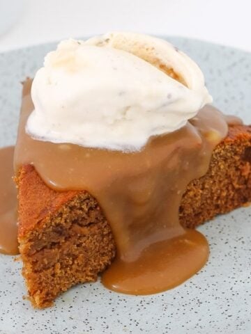 A slice of Sticky Date Pudding drizzled with caramel sauce and topped with vanilla ice cream on a plate.