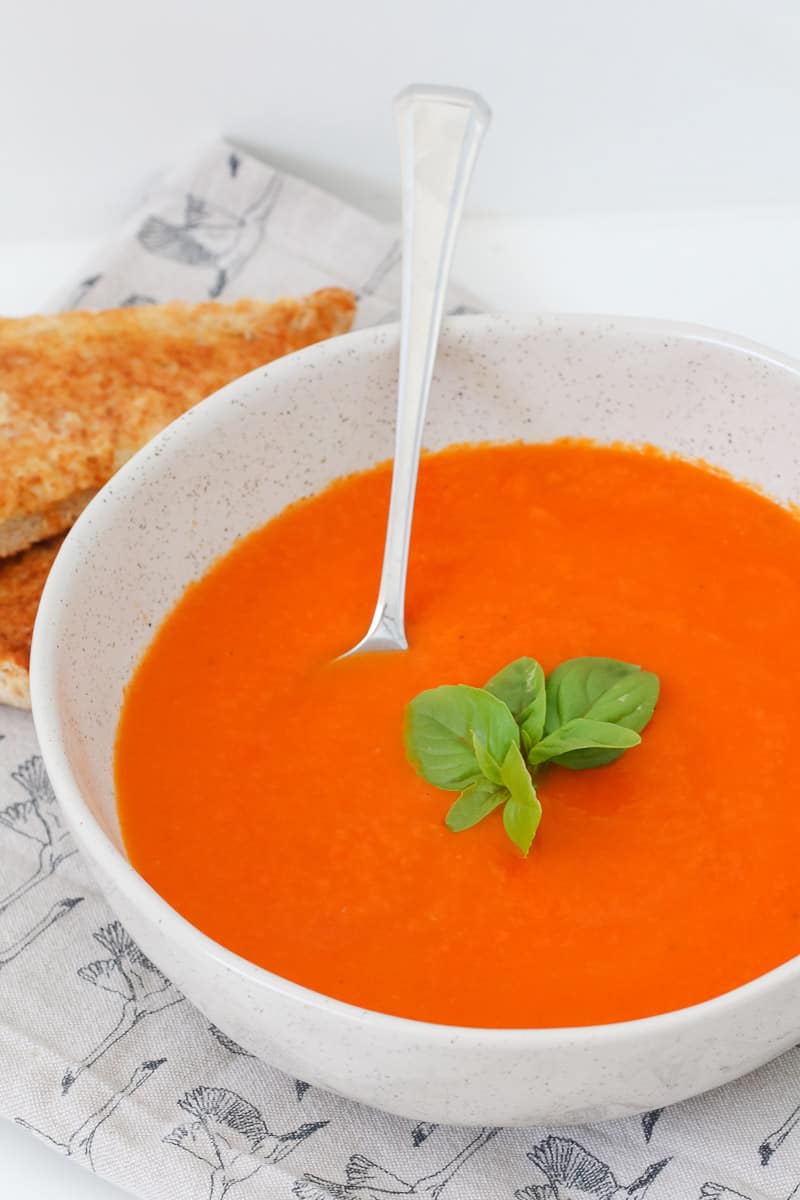 A bowl of tomato soup with fresh basil leaves and toast in the background.