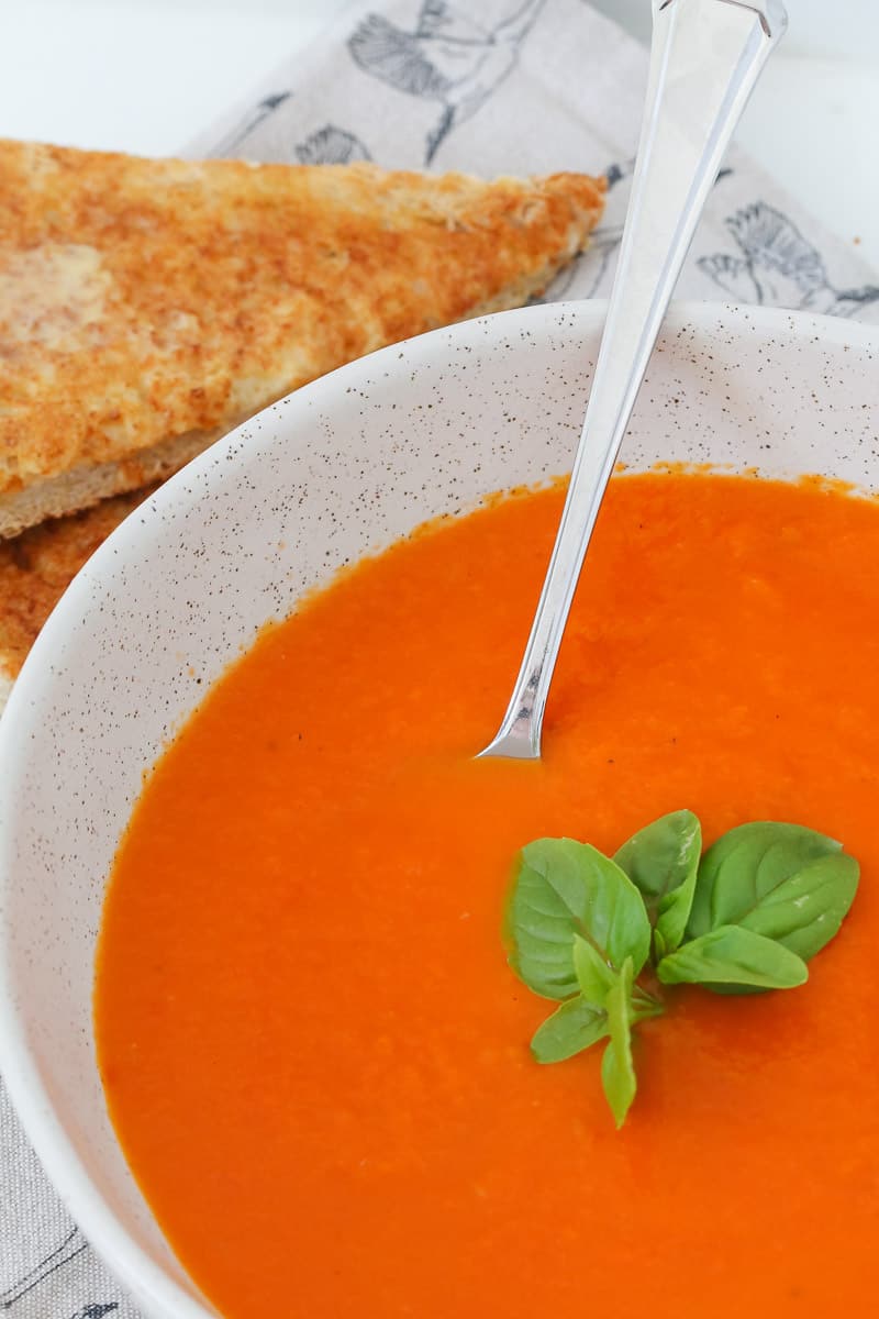 A bowl of red soup with fresh basil on top and buttered toast in the background.