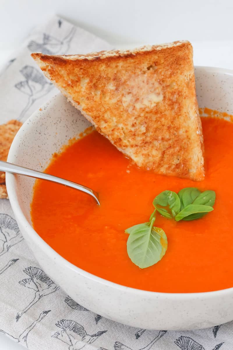 A pice of buttered toast being dipped into a bowl of tomato vegetable soup with fresh herbs on top.